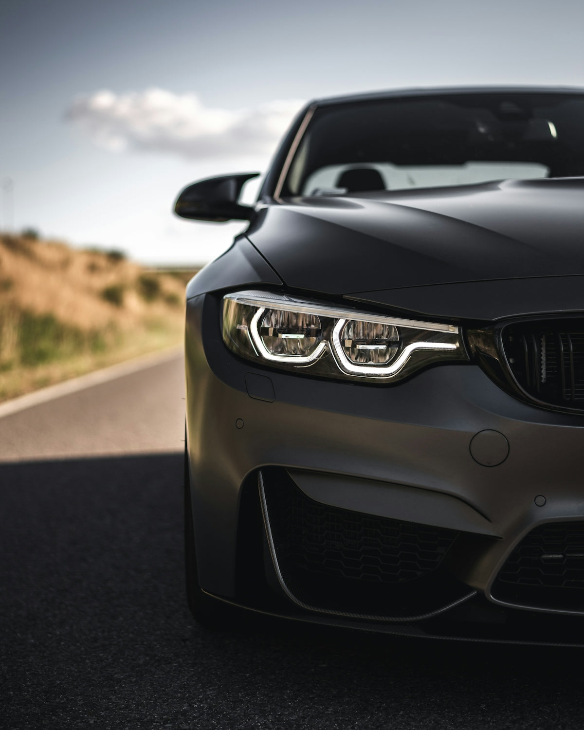A matte grey sports car with illuminated headlights on a paved road