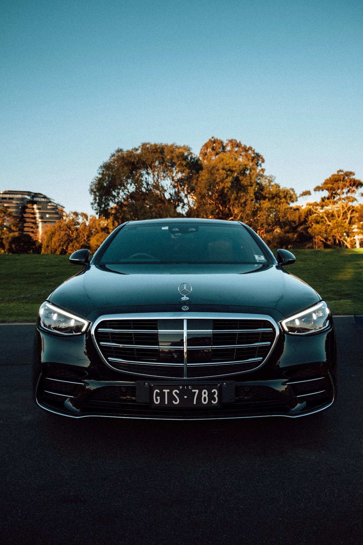 A black luxury sedan parked on asphalt before a grassy park at sunset.